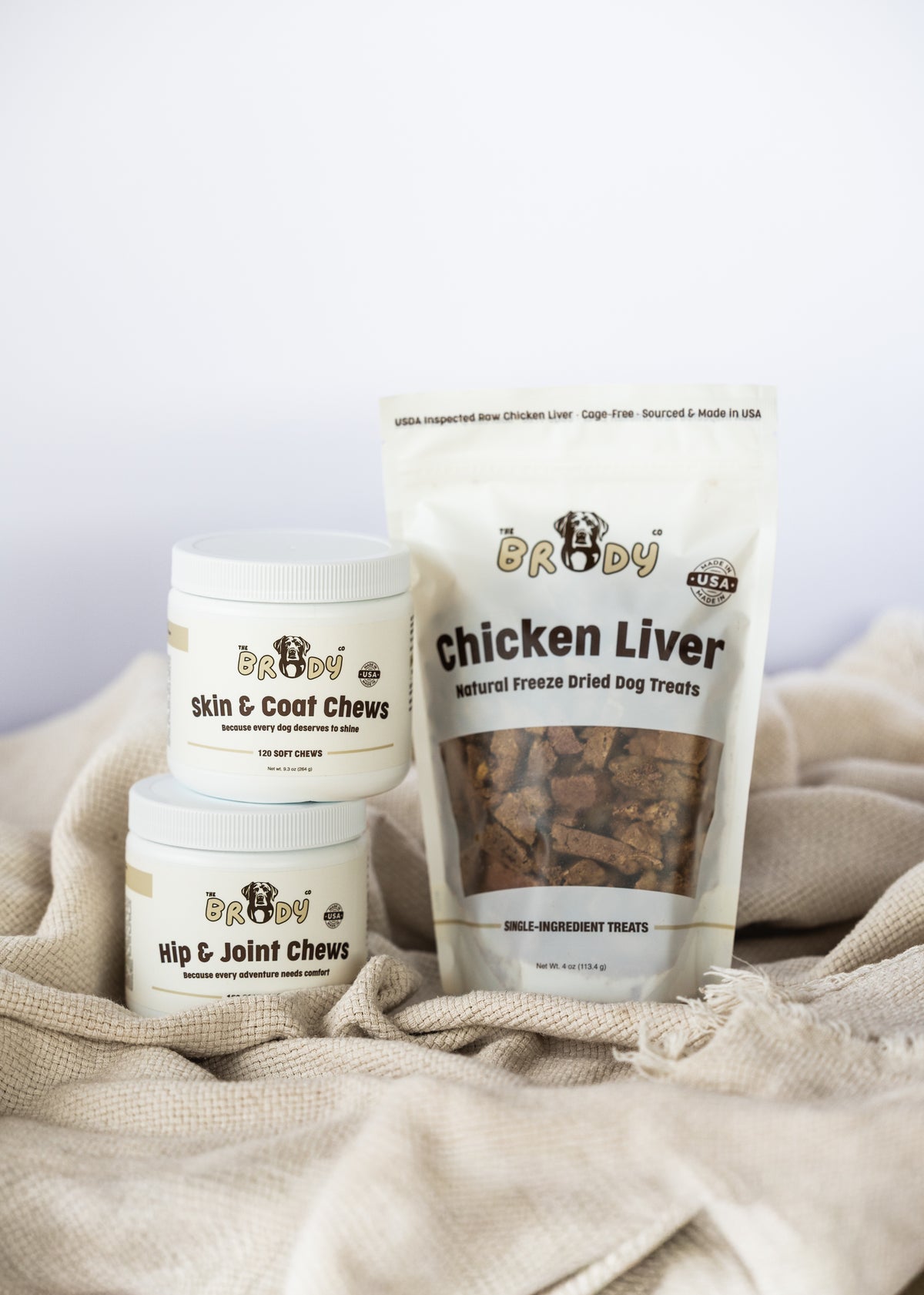Three Brody pet supplement products arranged on a beige fabric surface: two white jars labeled skin & coat chews and hip & joint chews, and one stand-up pouch labeled chicken liver natural freeze dried dog treats.