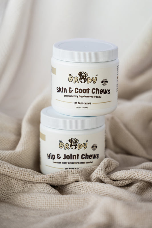 Two white plastic jars of dog chews stacked on a beige textured blanket. The top jar is labeled 'BRODY Skin & Coat Chews' and the bottom jar is labeled 'BRODY Hip & Joint Chews'.