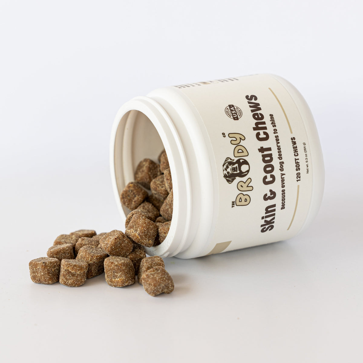 A white plastic jar of Brandy Skin & Coat Chews, tipped on its side with several brown kibble-style chews spilling out onto a clean white surface.