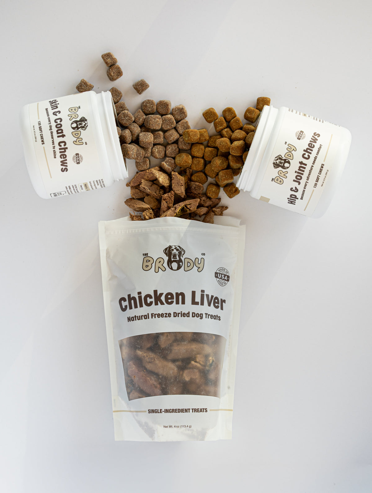 Three white containers of The Brody Co pet supplements and a large resealable pouch of Chicken Liver natural joint support treats, with some brown kibble chews spilled on a clean white background.